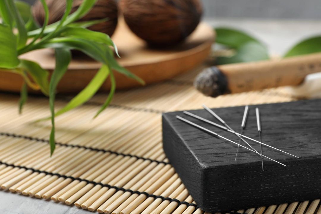 Black massage stone with silver pins on a bamboo mat, beside green leaves and wooden spa decor