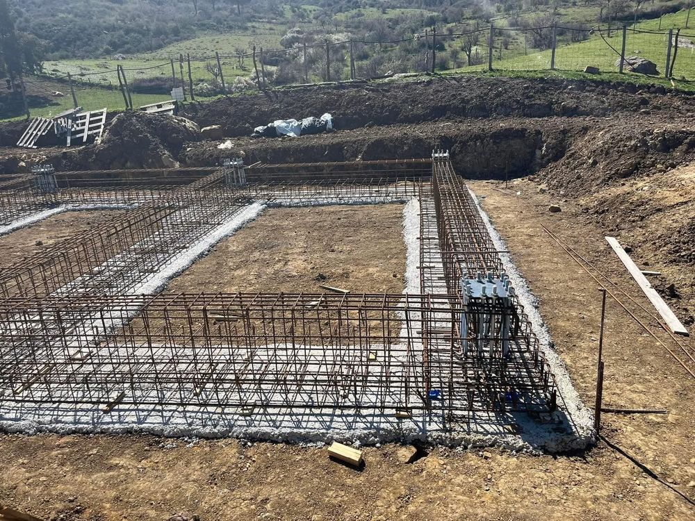 Cantiere con struttura in tondino di ferro e fondamenta in cemento; sullo sfondo il pendio della collina.