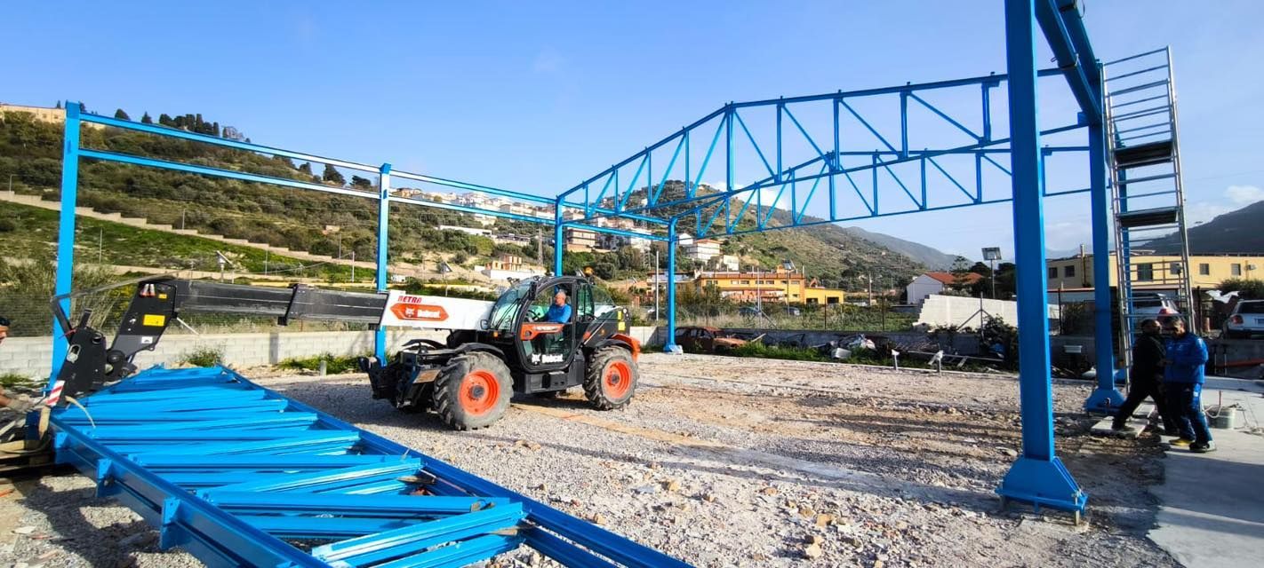 Un cantiere con una struttura metallica blu in fase di assemblaggio, con un carrello elevatore Bobcat e montagne sullo sfondo.
