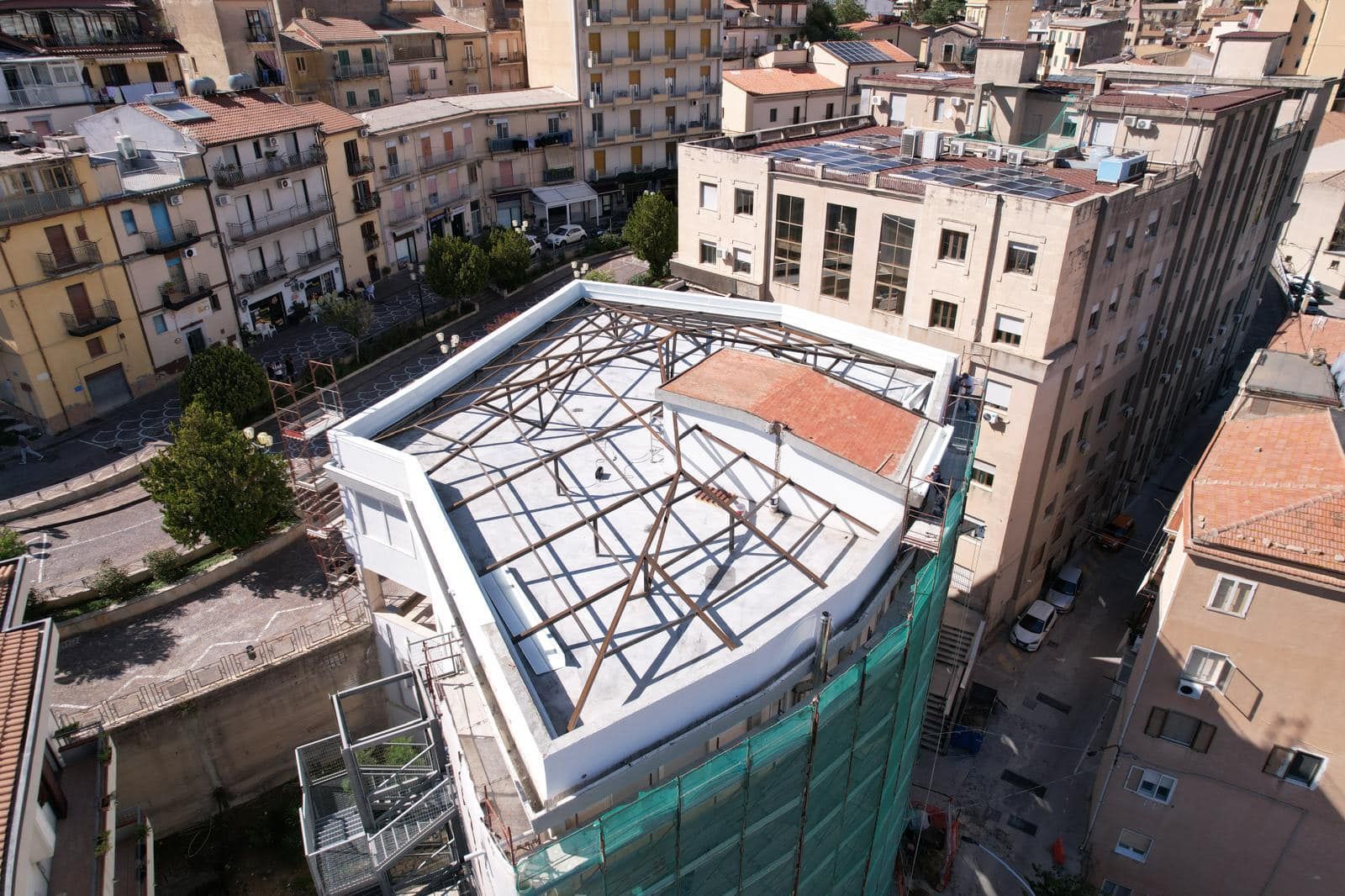 Vista aerea di un edificio in costruzione, che mostra un tetto con travi di sostegno. Edifici circostanti in un contesto urbano.