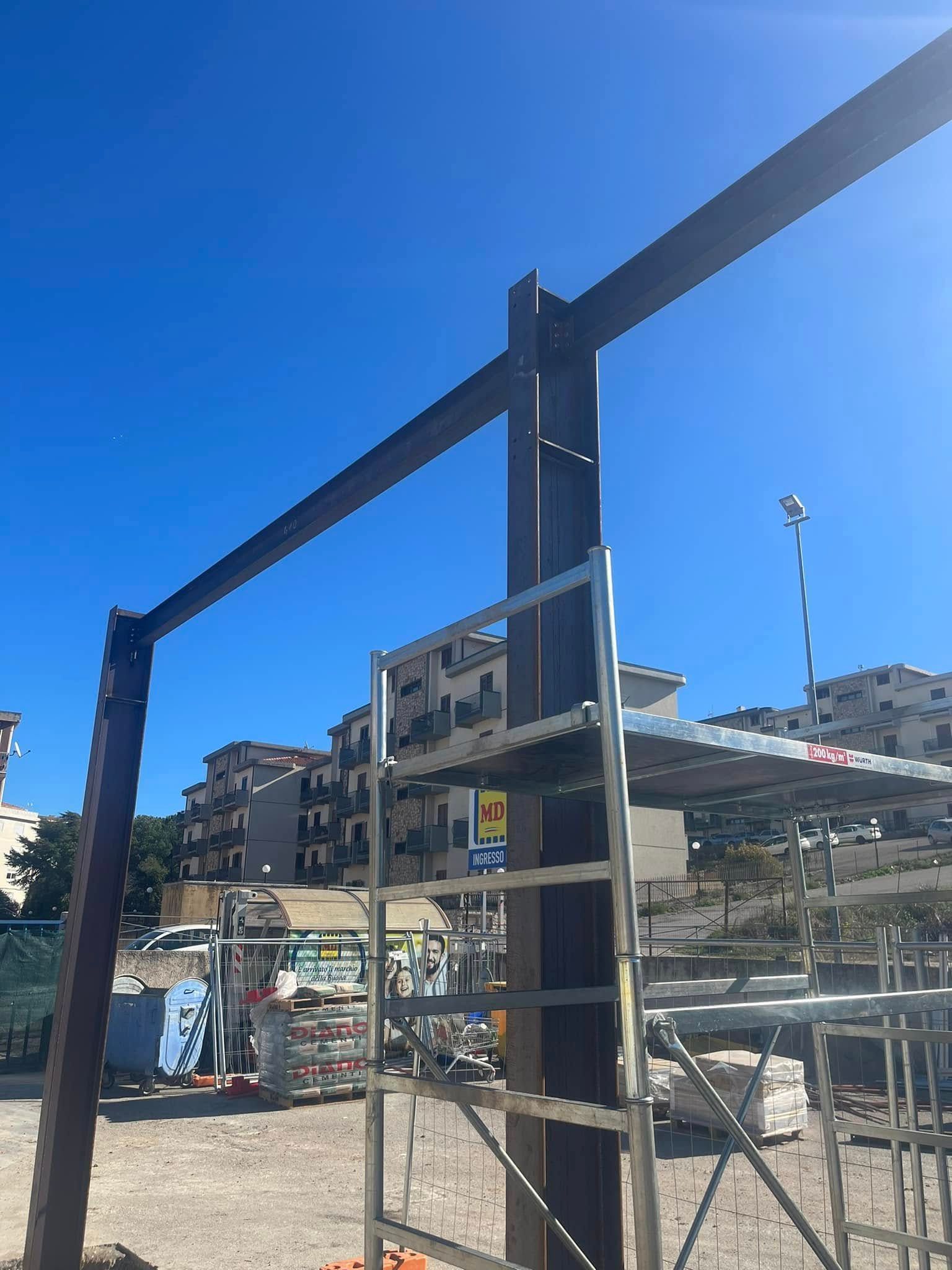 Travi d'acciaio sotto un cielo terso e azzurro, parte di un progetto di costruzione. Una scala è appoggiata a una trave di sostegno.