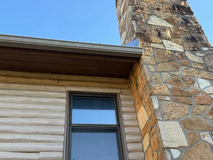 A side view of a stone chimney meeting a house wall with horizontal siding, eaves, and a window under a clear blue sky.