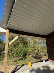 An outdoor porch view featuring a wooden support post, white vinyl soffit ceiling with recessed lighting, and brick wall.