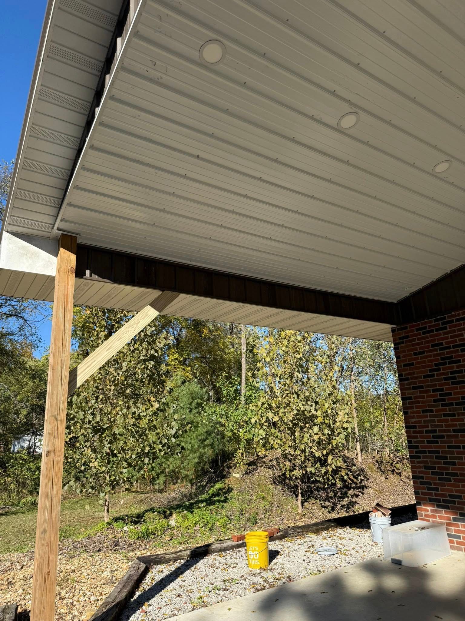 A low-angle view of a patio ceiling with white soffit panels, recessed lights, a wooden support post, and a brick wall.