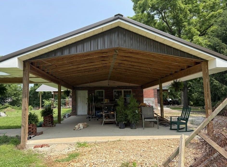 A covered patio with a wooden ceiling, concrete floor, furniture, and plants attached to a brick house on a sunny day.
