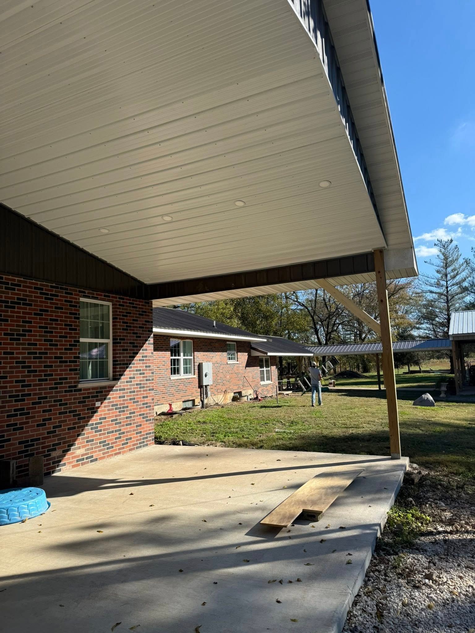 A concrete patio under a covered porch attached to a red brick house, with a person standing in the background yard.