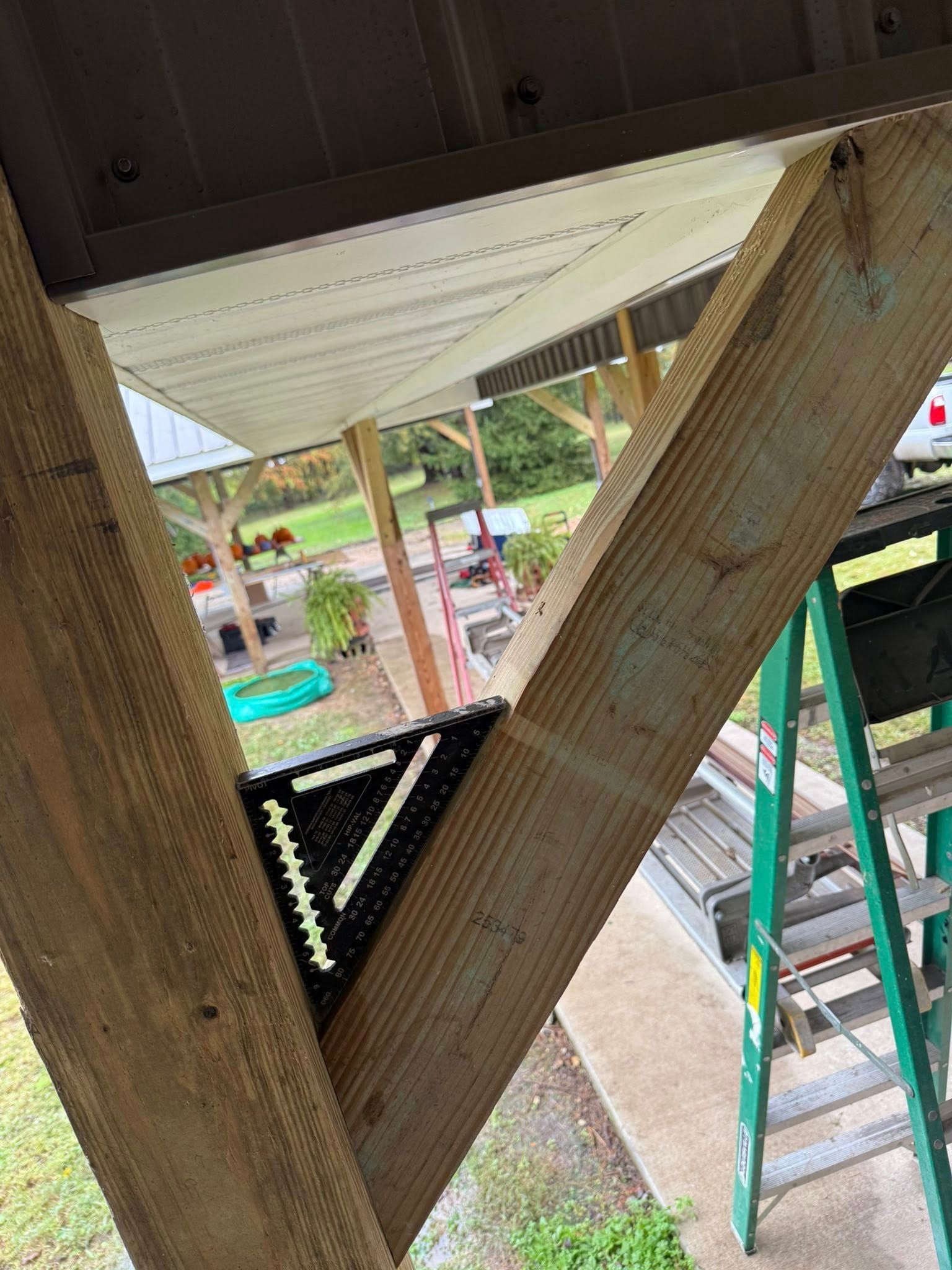 A speed square rests on a wooden support beam angled against a vertical post, with a ladder visible in the background.