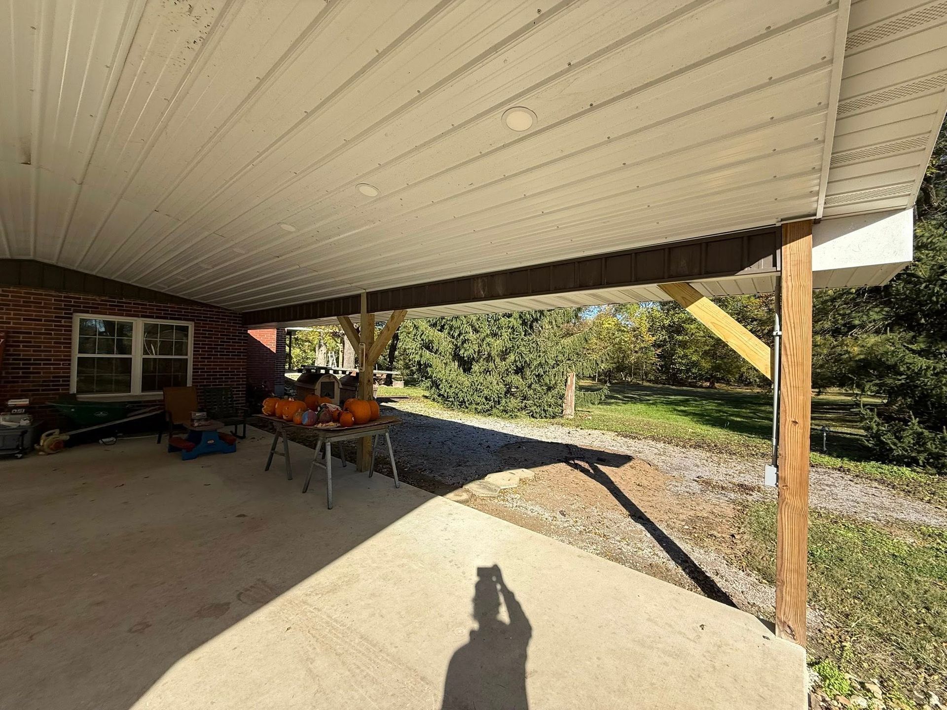 An outdoor covered patio area with a concrete floor, wooden support beams, and a view of trees and grass in the distance.