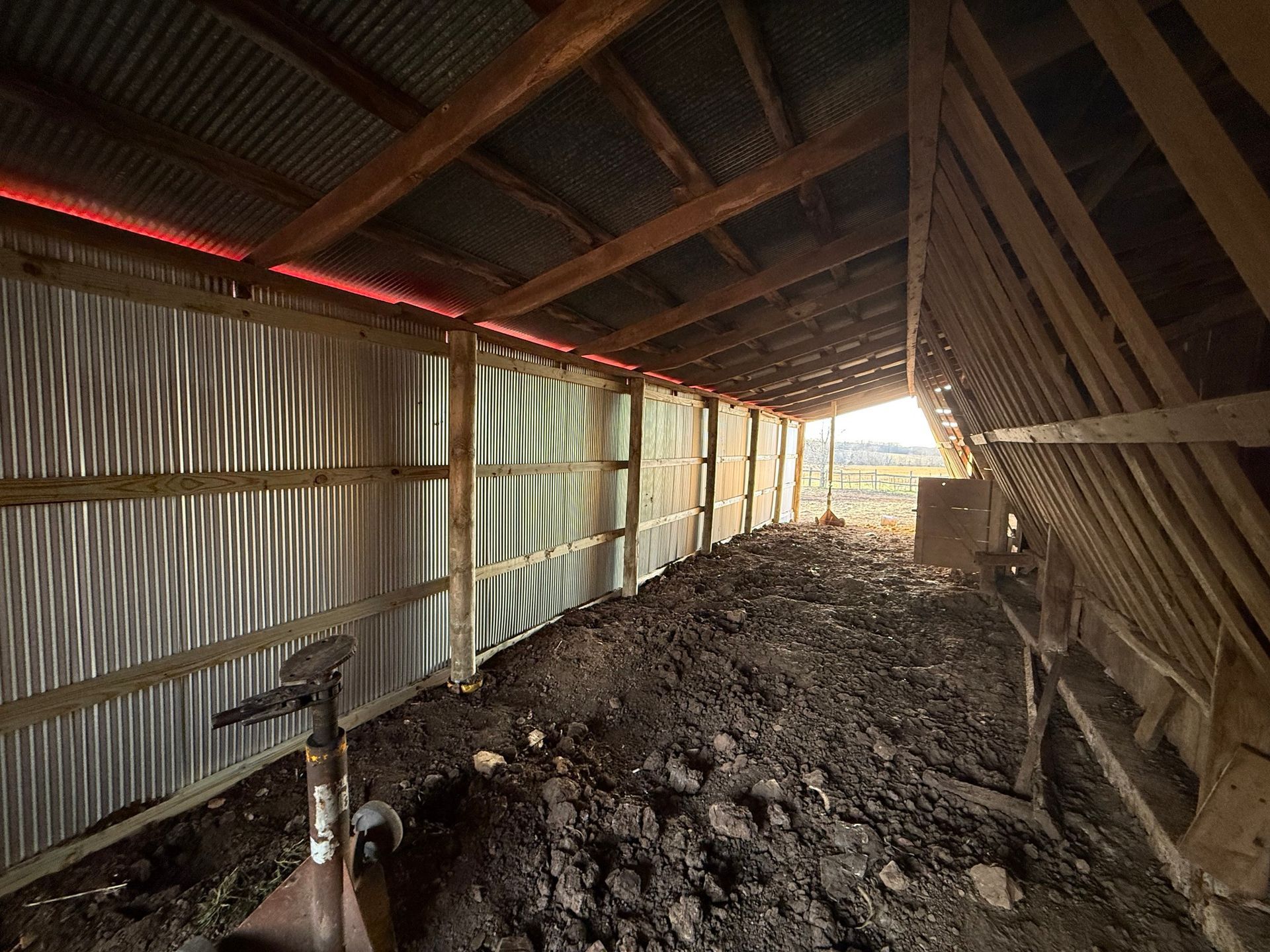 The interior of a shed with a corrugated metal wall, a wooden frame, and a muddy, dirt-covered floor.