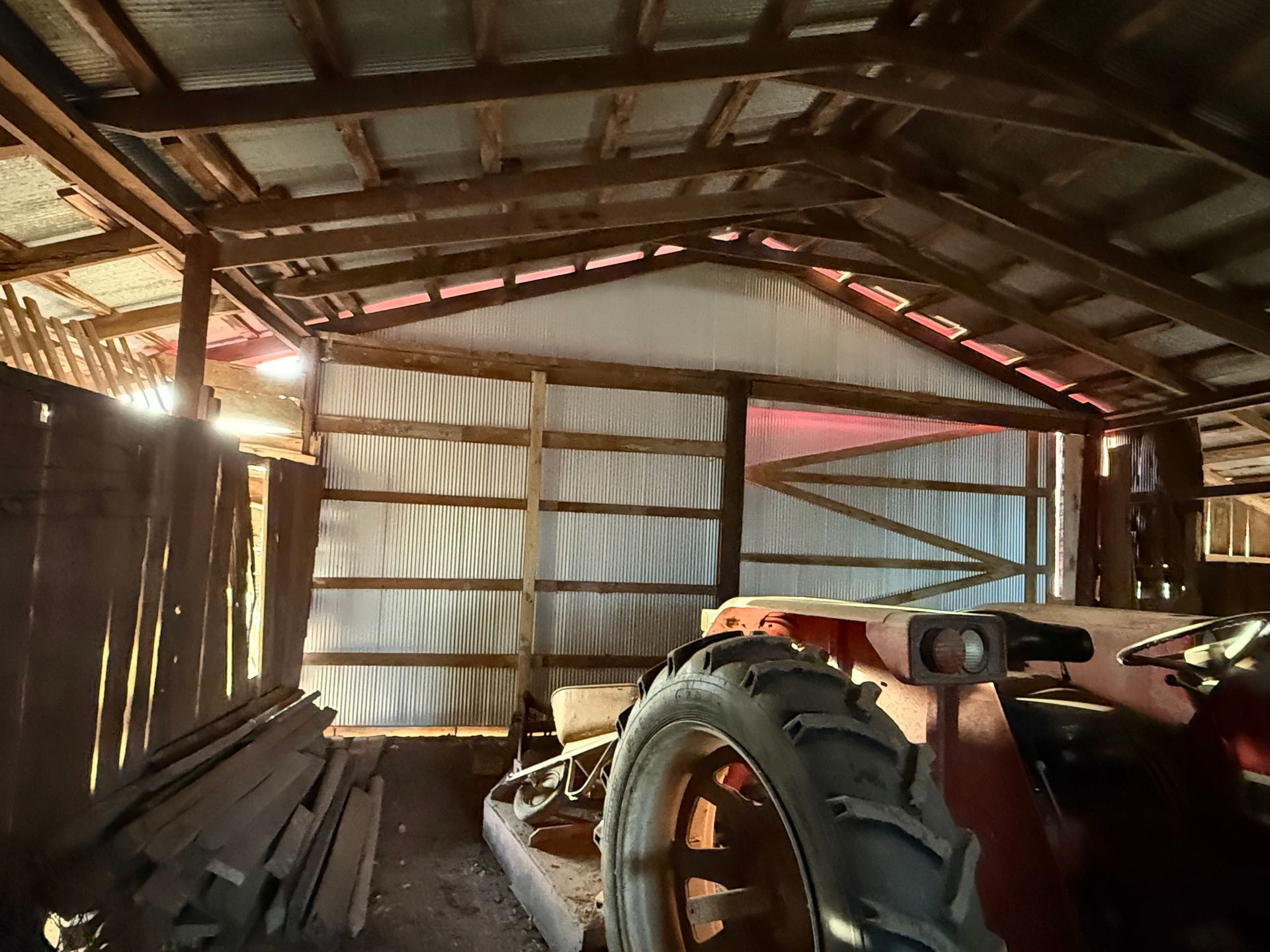 An old red tractor parked inside a rustic barn with metal walls and exposed wooden rafters.