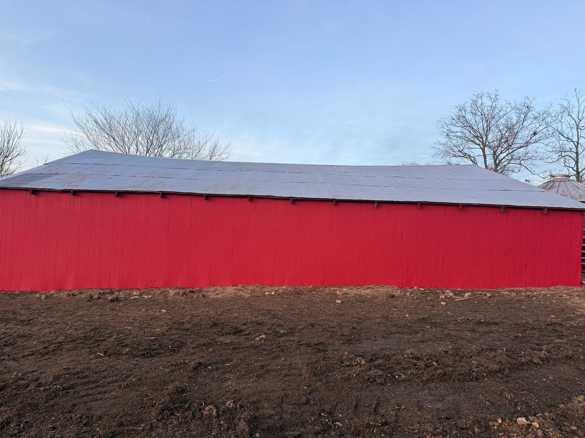A long, rectangular building with a red-pleated fabric wall and a grey metal roof, situated on a plot of brown soil.