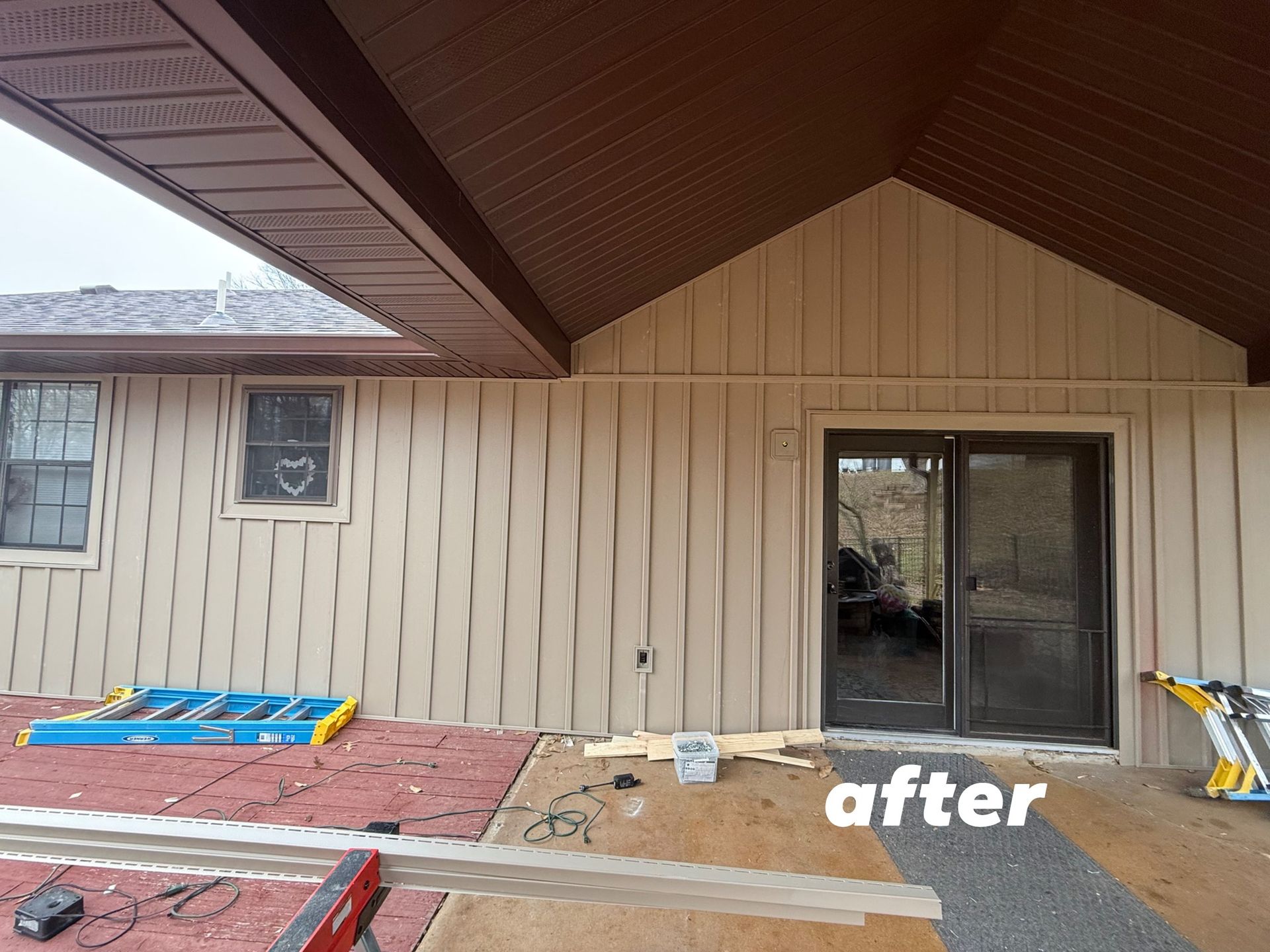 A tan-sided house exterior with a brown roof and a large glass sliding door, seen during a patio renovation.