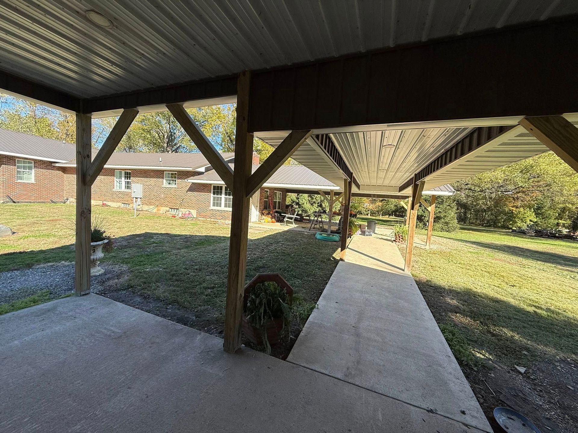 A concrete walkway under a long covered patio structure connects a brick house to a yard on a sunny day.