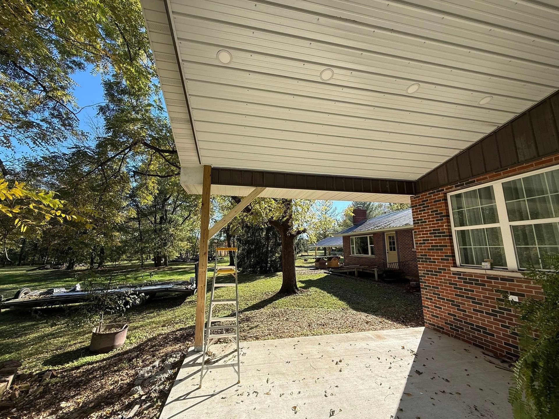 A patio with a white soffit ceiling, a wooden support post, a step ladder, and a brick house wall under a clear blue sky.