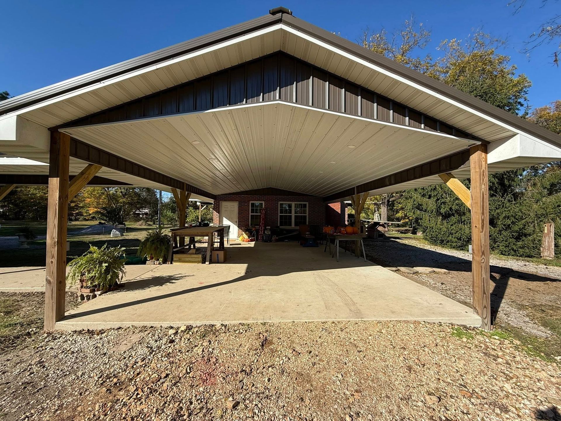 A large, open-sided wooden carport with a metal roof shelters a concrete patio area in a sunny, tree-lined setting.
