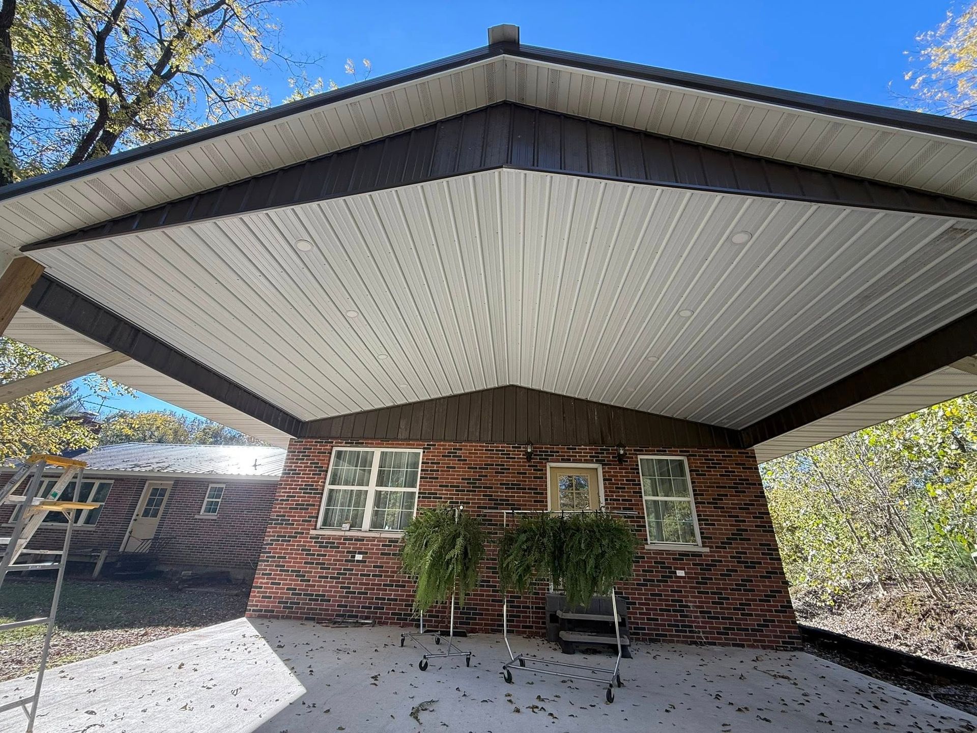 A brick house exterior with a large, white-paneled covered porch, a concrete patio, and nearby trees under a blue sky.
