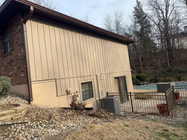 Tan vertical siding on the exterior of a brick home with a small window, an HVAC unit, and a metal fence in the yard.