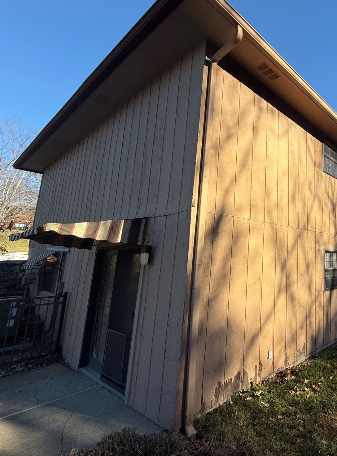 A tan two-story building exterior with vertical siding, a small awning over a glass sliding door, and grass in the yard.