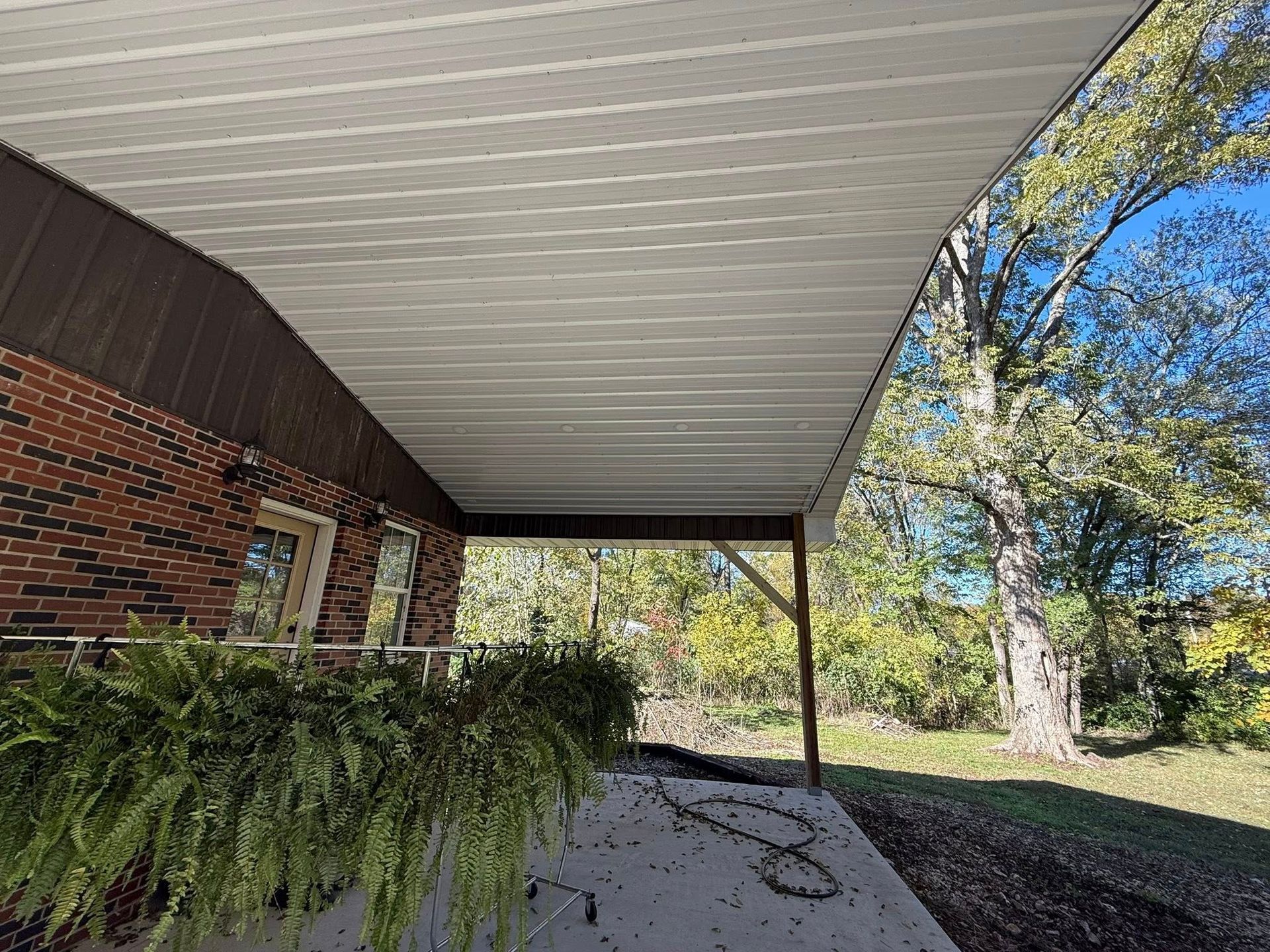 A covered concrete patio attached to a red brick house, with a large fern in the foreground and trees in the background.