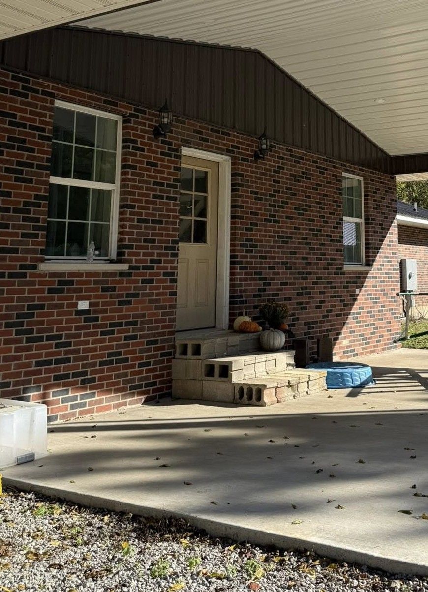 Brick house exterior with a covered concrete patio, a light-colored door, concrete block steps, and two windows.