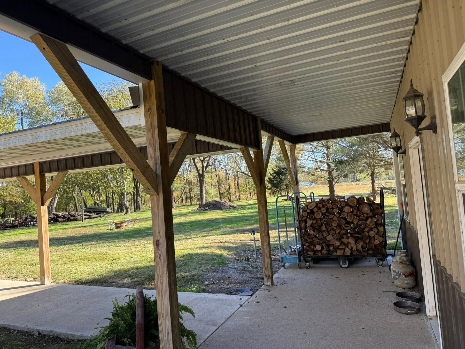 A concrete porch with wood-post support beams, metal roof, and a firewood rack, overlooking a sunny, grassy field.