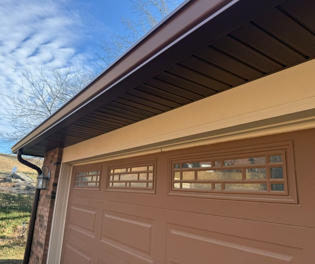A brown garage door with rectangular window inserts, topped by brown soffit and trim against a bright blue sky.