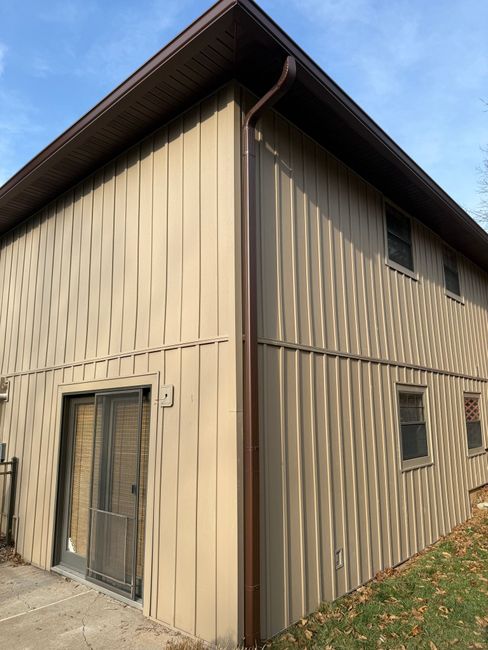 A two-story building with tan vertical siding, brown trim, a sliding glass door, and shadows cast across the walls.