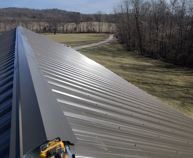 A dark brown metal roof with a ridge cap being installed, overlooking a grassy field and trees.