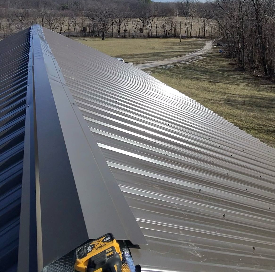 A view looking down the peak of a dark metal roof during installation, with a power drill resting on the metal ridge cap.