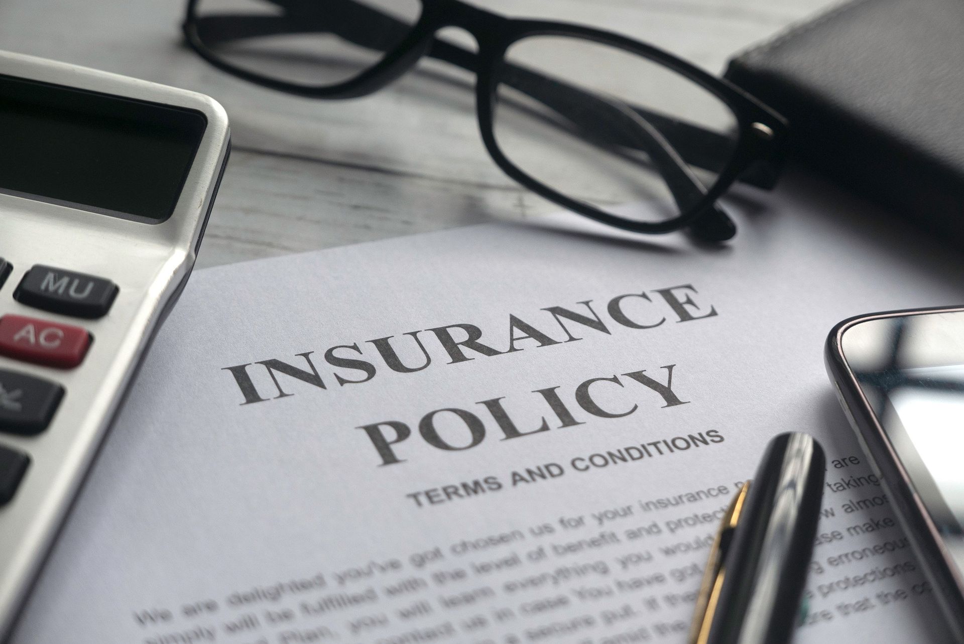A calculator , glasses , a pen , and a cell phone are on top of an insurance policy.