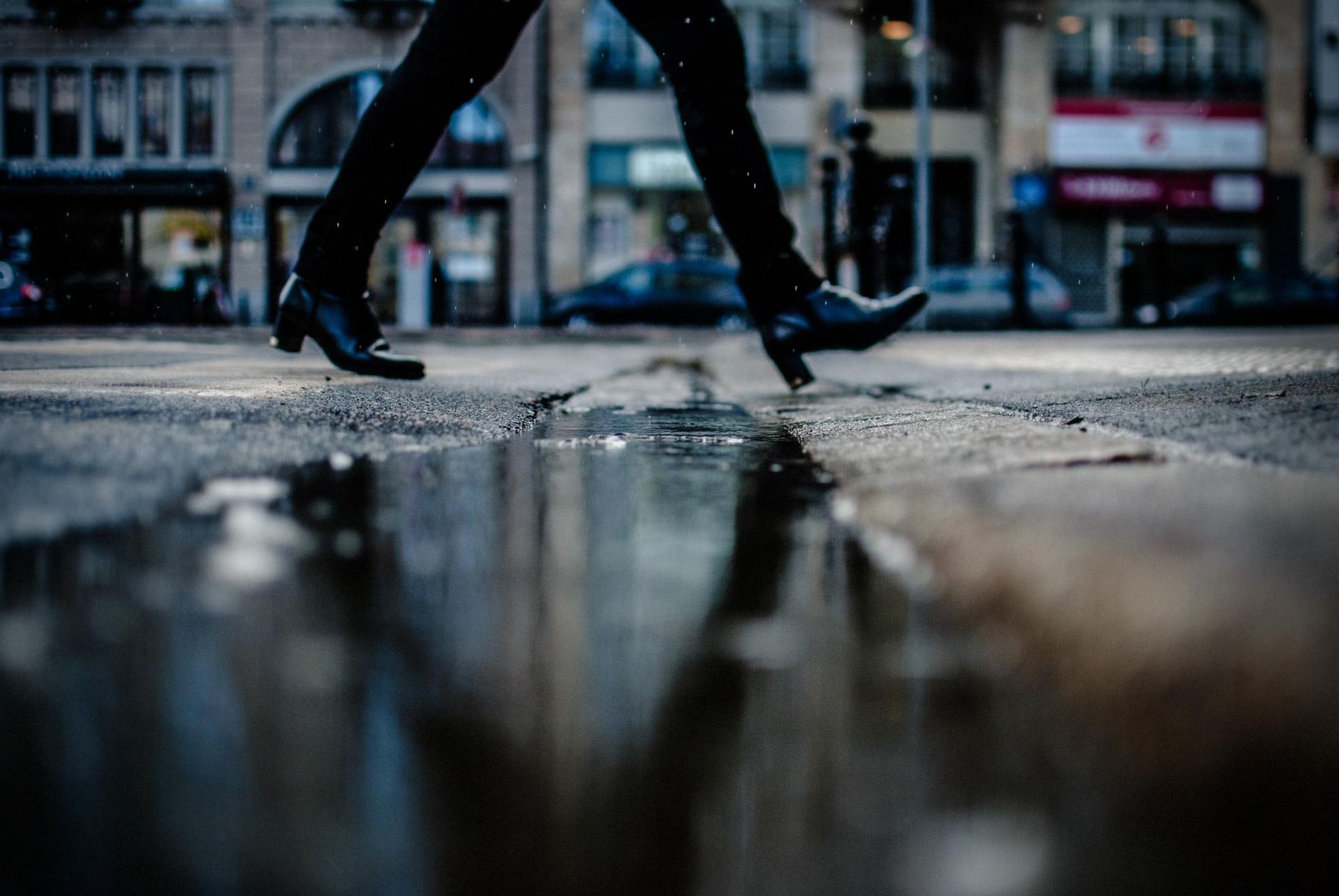 Person's legs in black boots walking across a wet street, reflecting buildings and sky.