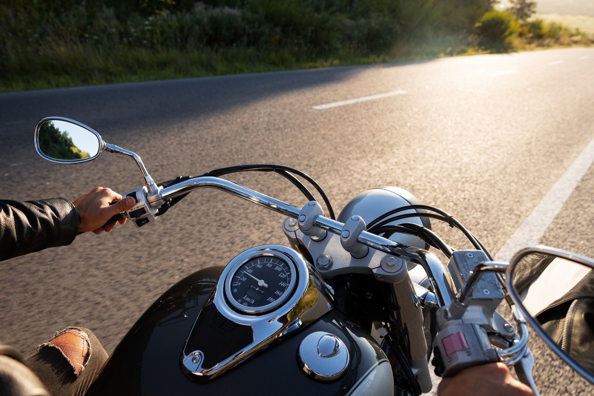 Motorcycle handlebars, gauges, and rider's hands with open road in the background. Sunny day.
