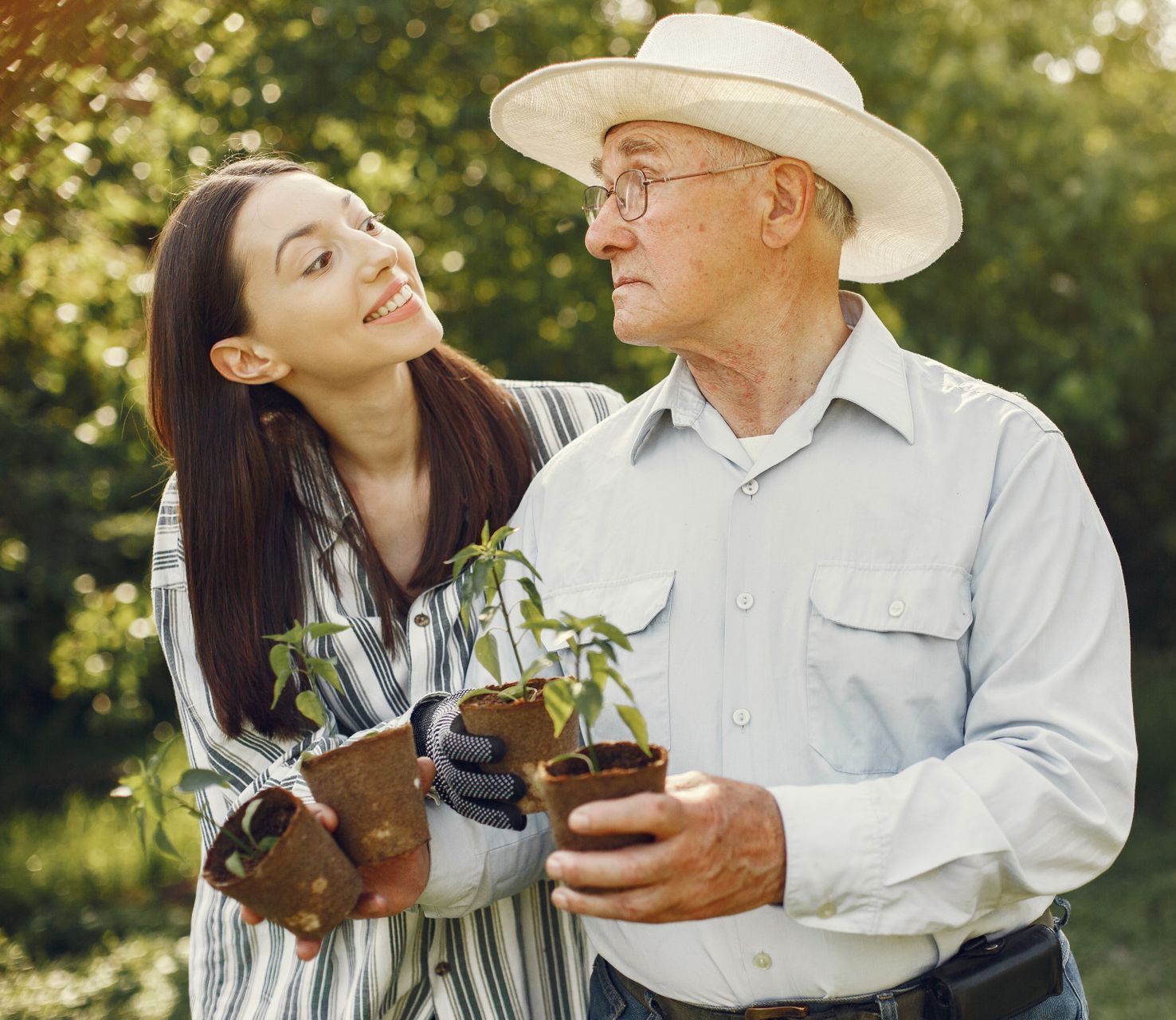 Woman and man, outdoors in garden, holding small plants in pots. Woman smiles, man wears hat.