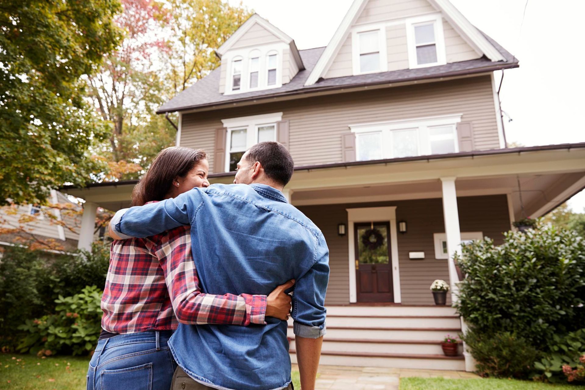 Couple with arms around each other looking at a house with a porch; a brown exterior and autumn foliage.