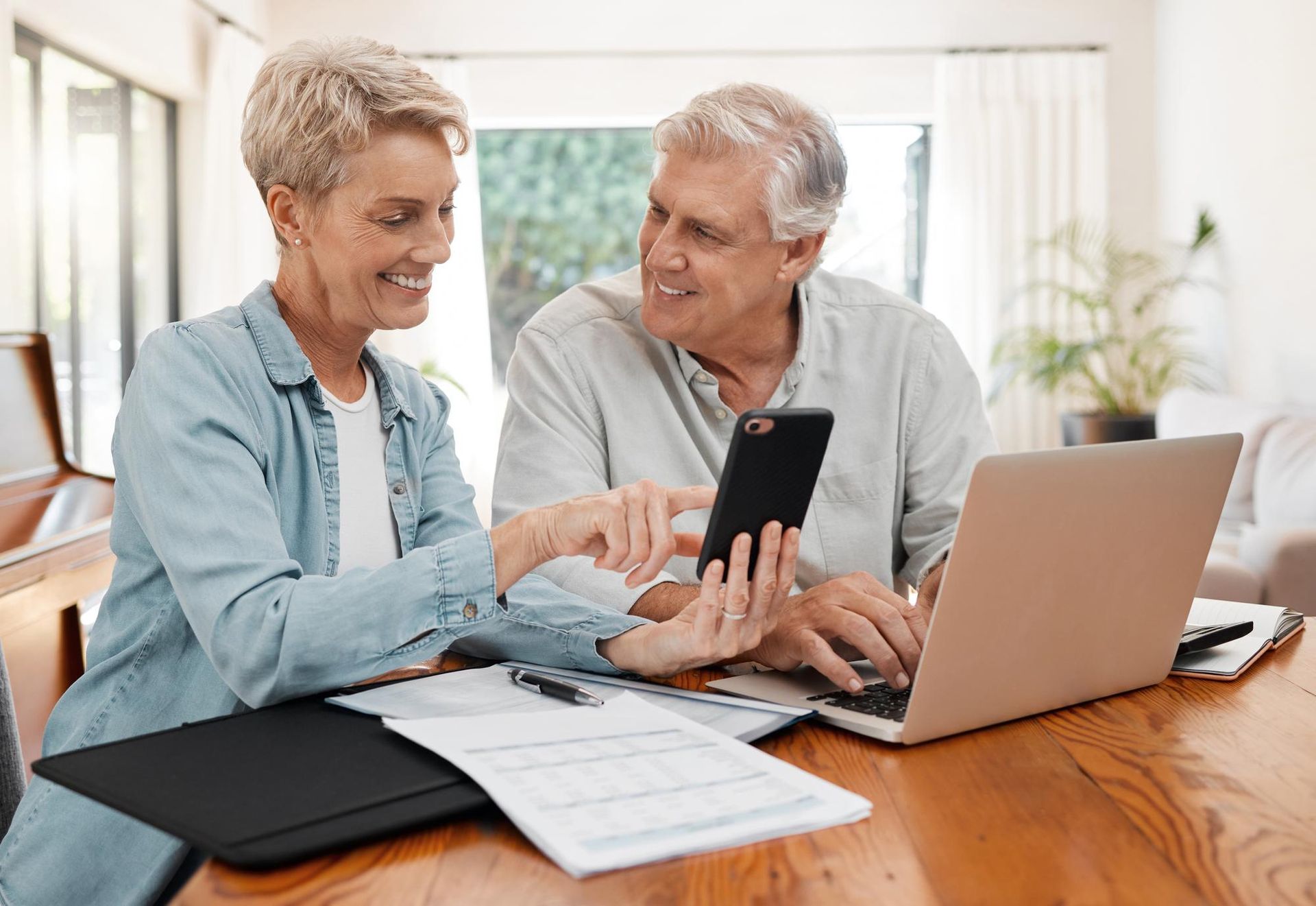 Older couple looking at a phone and laptop at a table, smiling. Documents and pen visible.