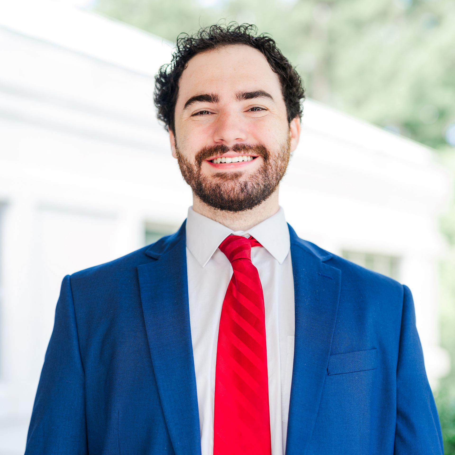 Man in blue suit, smiling, red and blue striped tie, brown curly hair, and beard.