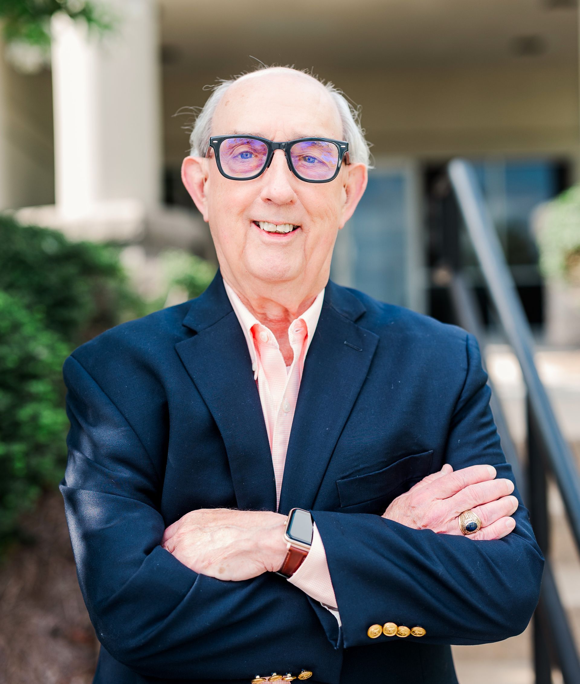Man in suit and tie, wearing glasses, smiling slightly, against a brown background.