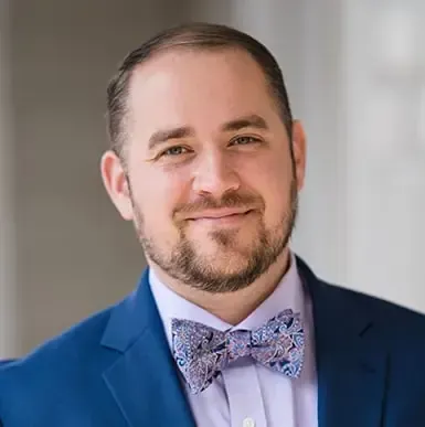 Man with brown hair and beard, wearing a patterned bow tie and blue suit, smiles.