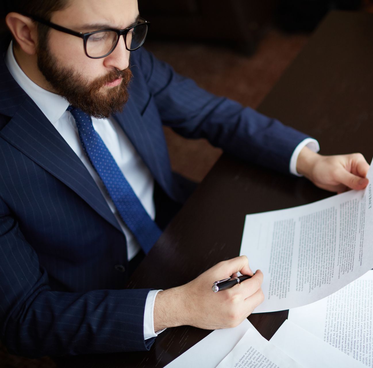 Man in suit with glasses reviewing documents, pen in hand.