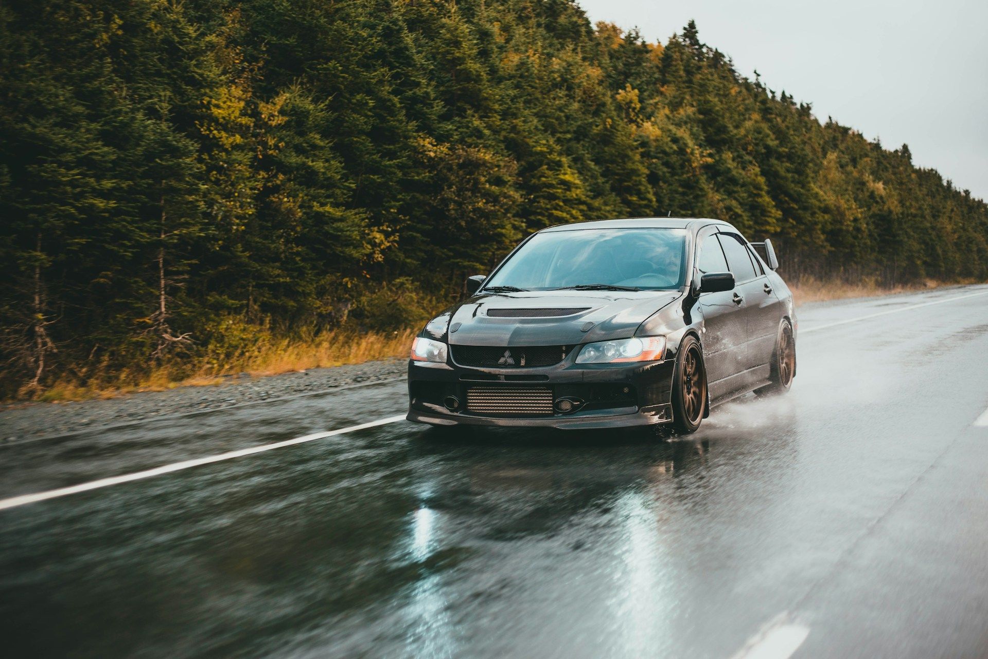 Black Mitsubishi Evolution car driving on wet road, trees in the background.
