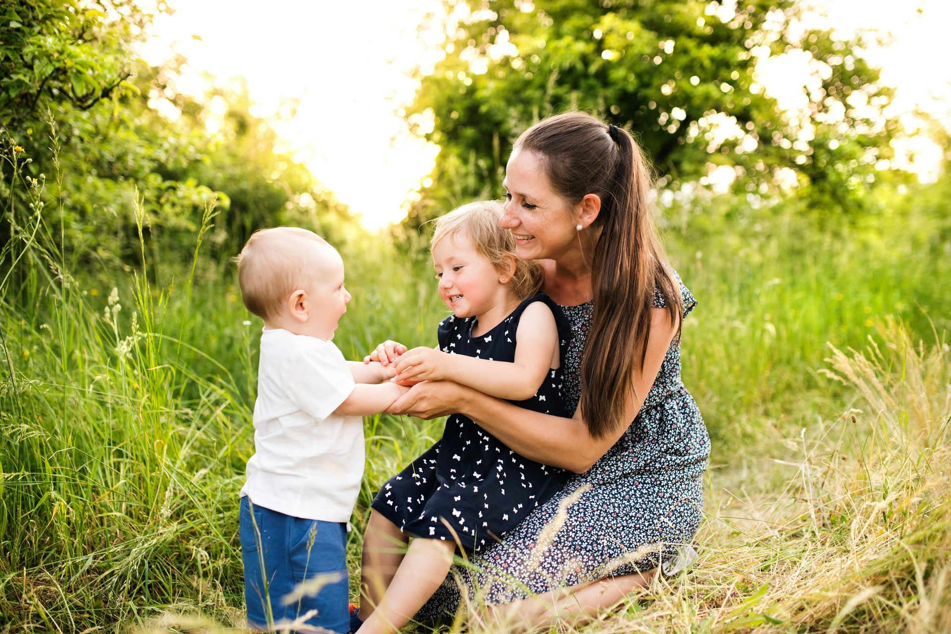 Woman with two young children smiling outdoors in tall grass.