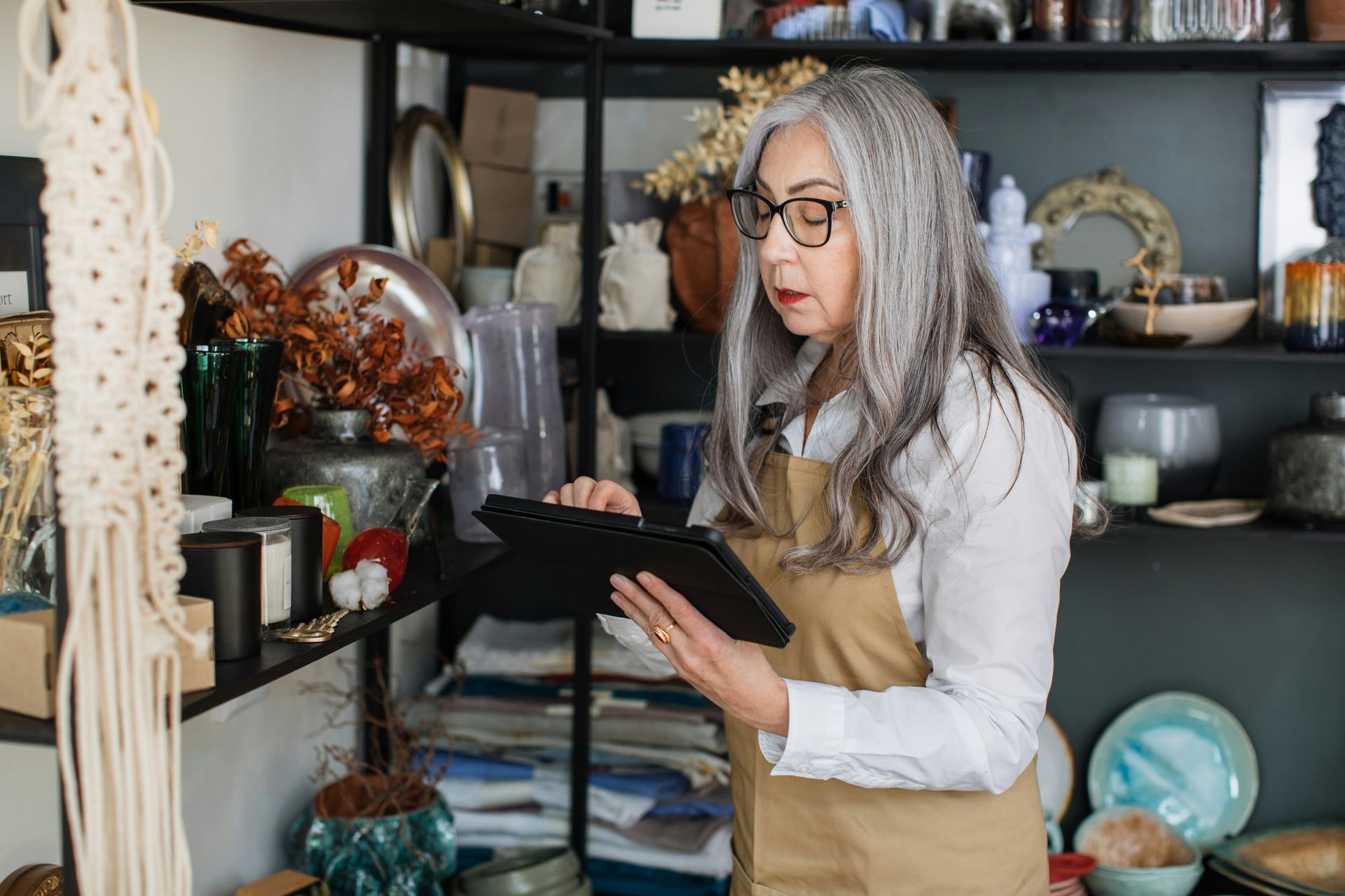 Woman in apron uses a tablet in a shop filled with crafts and decor.