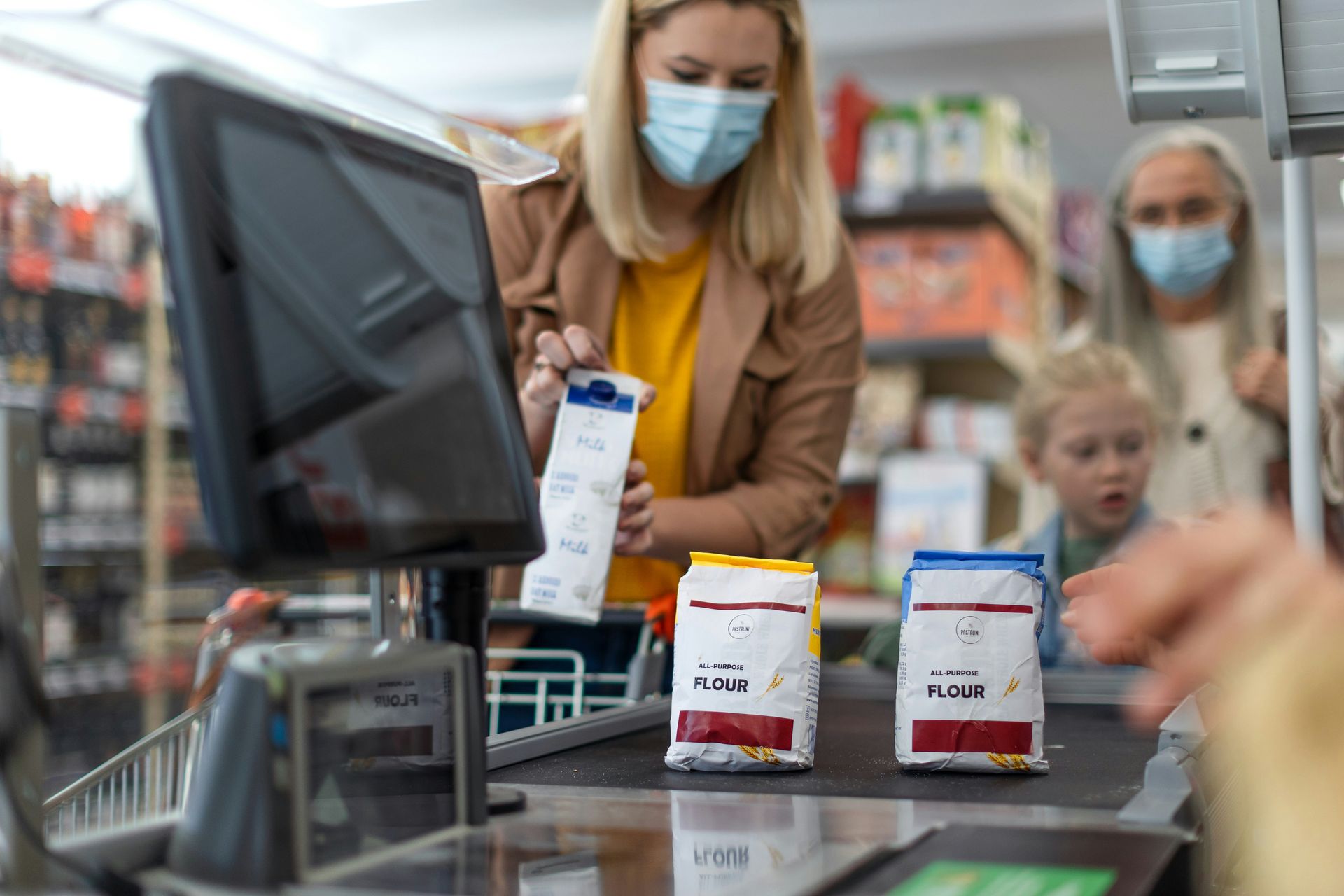 Woman at grocery store checkout, wearing a mask, scanning flour packages. Child and another person are nearby.