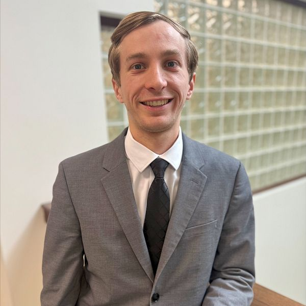 Man in gray suit and black tie smiles, standing in front of glass block wall.