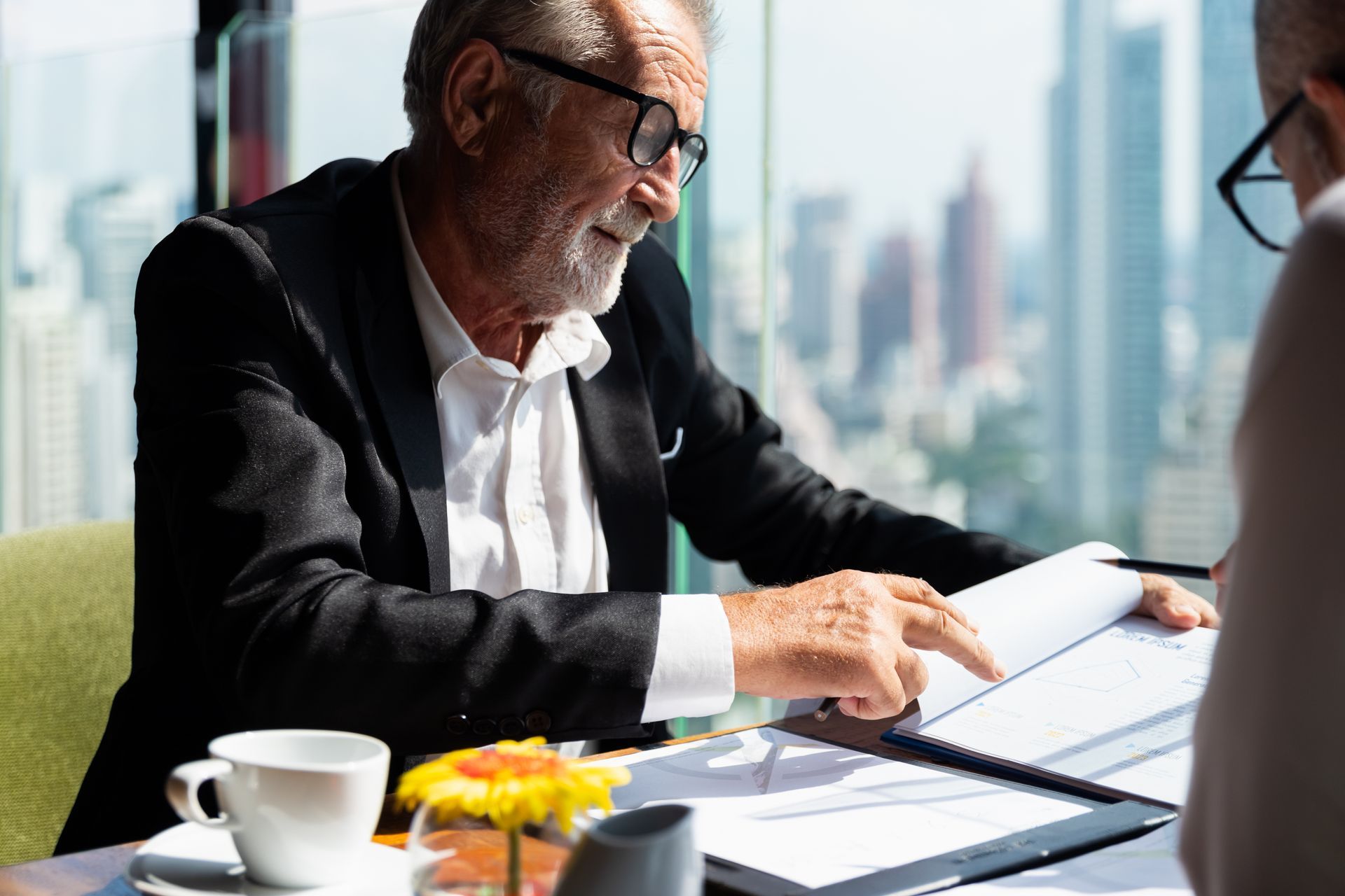 Man in suit points to papers during a meeting in a room overlooking a city.