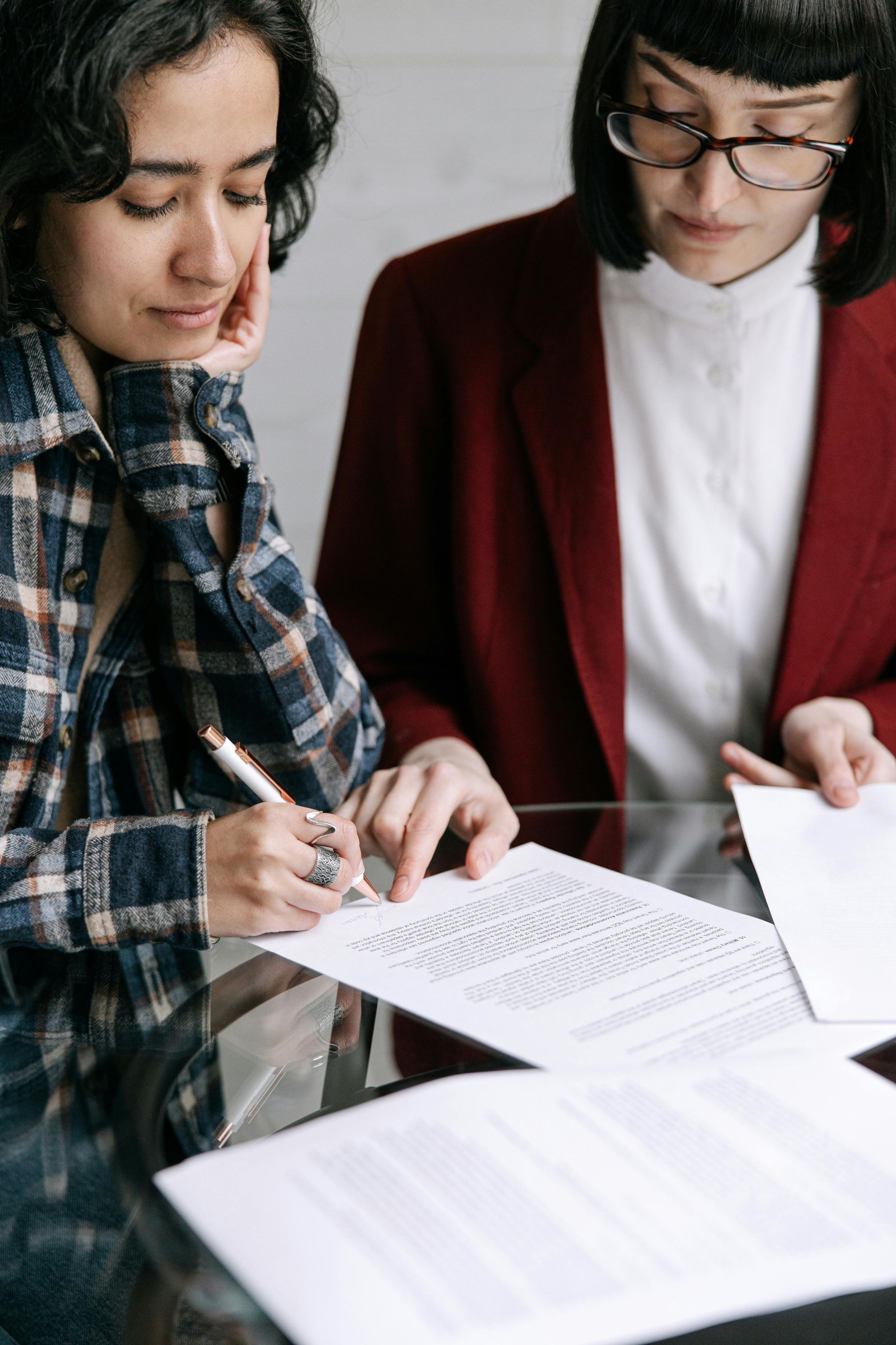 Two people reviewing and signing a document at a glass table. One wears a plaid shirt, the other a red blazer and glasses.