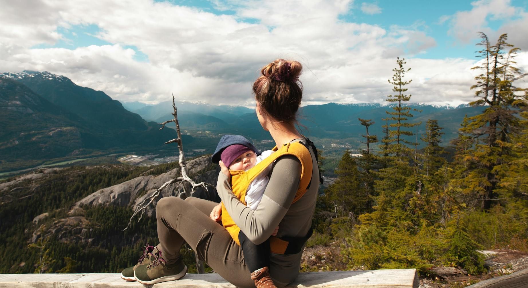 Woman with baby in carrier overlooks a mountain vista on a cloudy day.