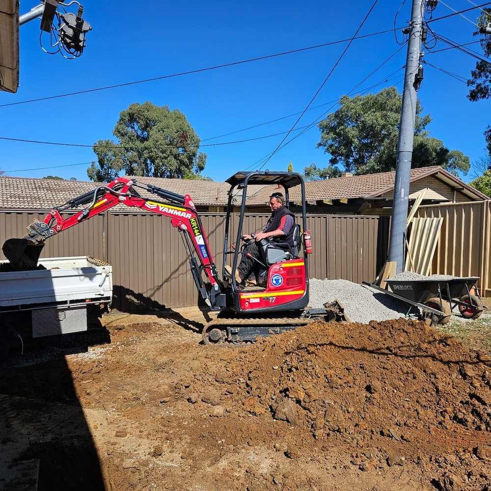 A Man Is Driving A Small Excavator On A Dirt Field — Around Town Plumbing Services in Canberra, ACT