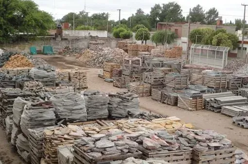 Montones de losas de piedra gris y ladrillos apilados en un patio exterior con árboles y casas al fondo.