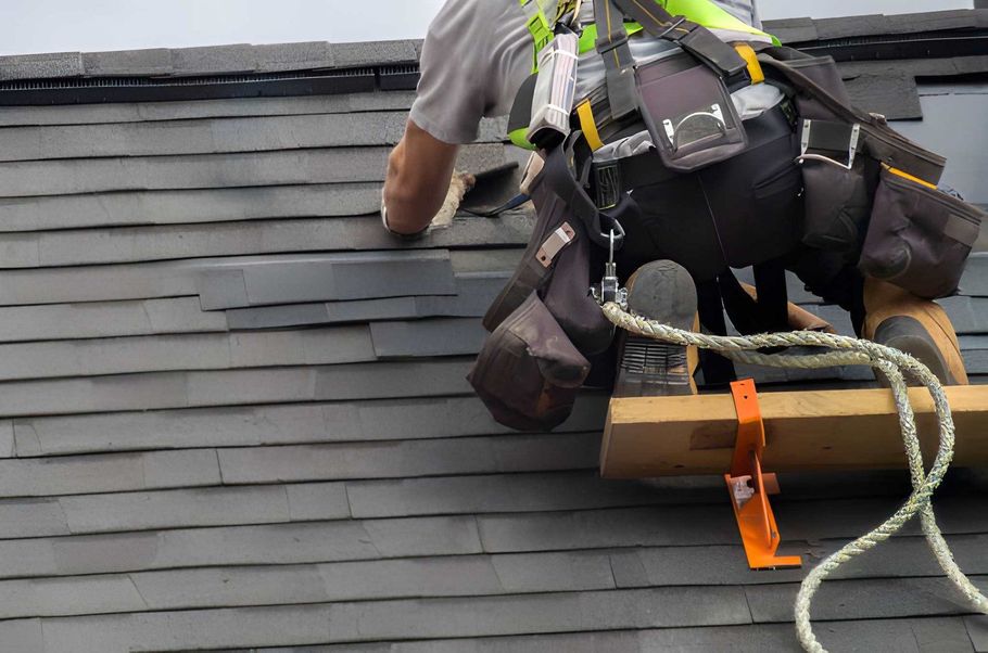 Worker on a roof installing gray shingles, wearing a safety harness and kneeling beside tools and rope.